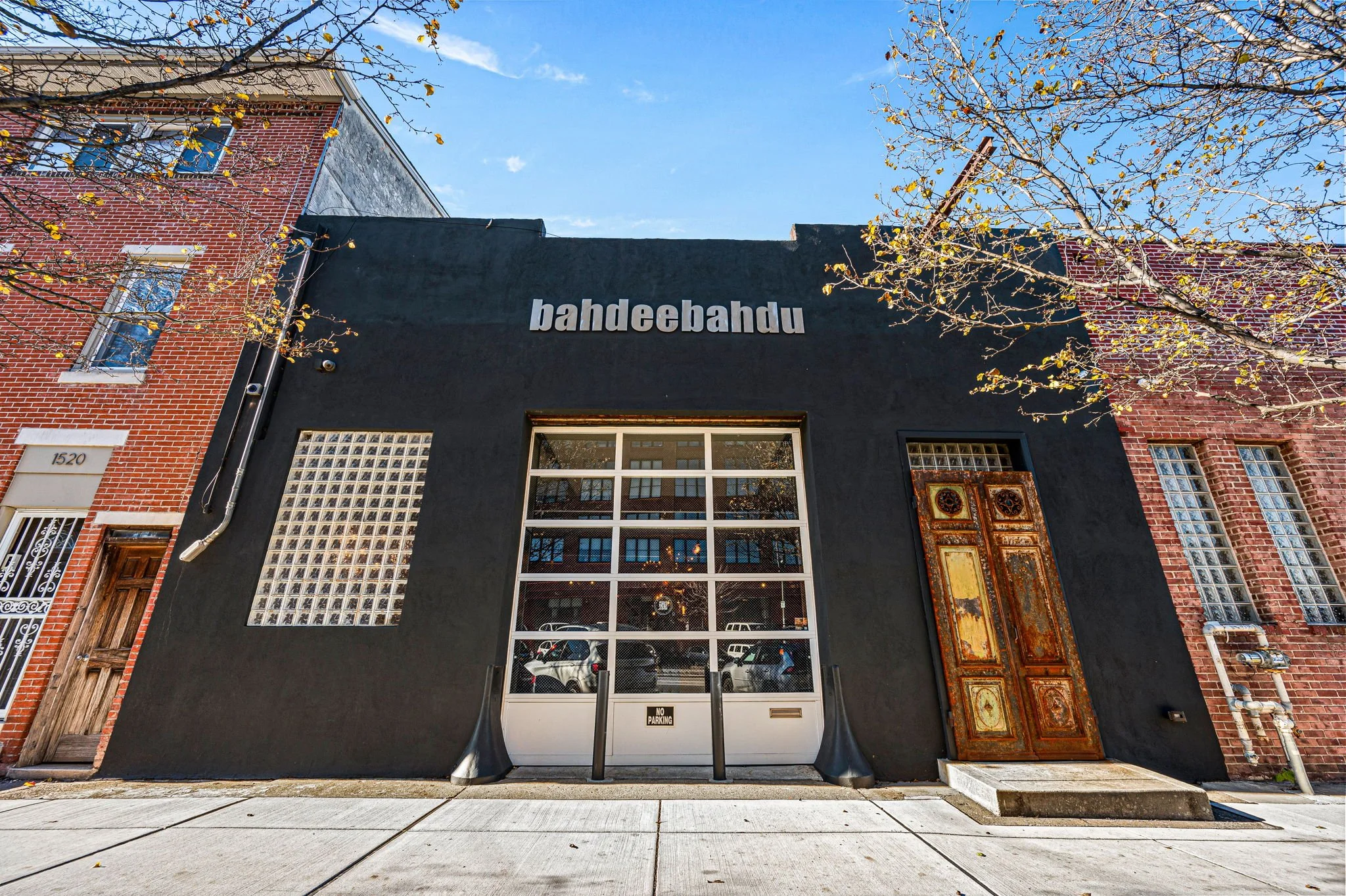 A black storefront with the sign 'bahdeebahdu' above the entrance. The building has a large window and an ornate, rusted door to the right. There are trees with yellowing leaves and brick buildings on either side. The sky is blue with some clouds.
