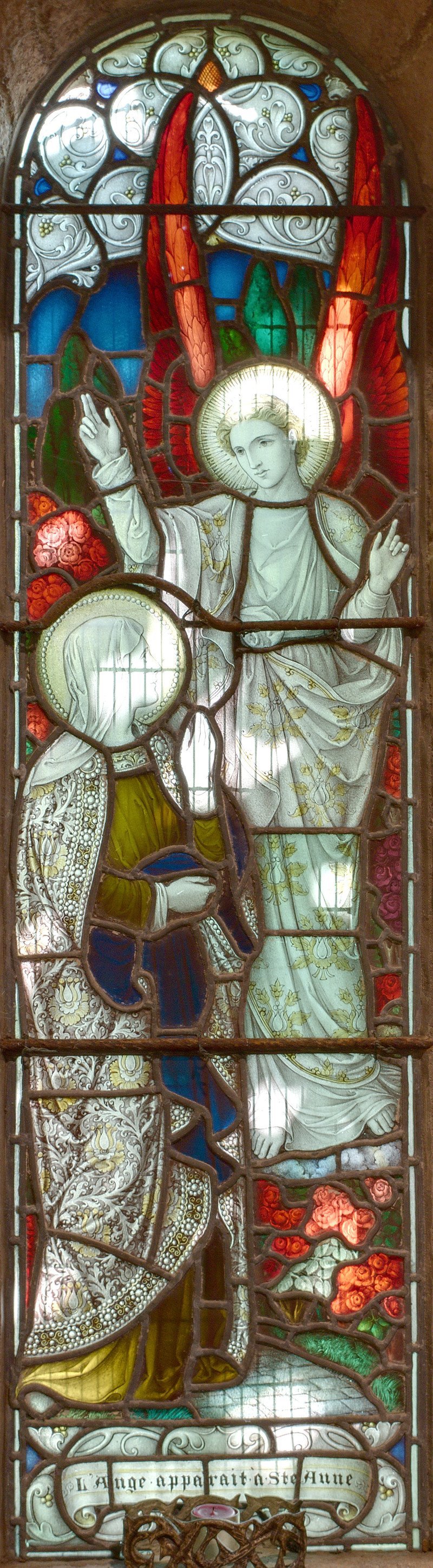 A stained glass window depicting the Virgin Mary and a saintly figure, possibly an angel or Jesus, with a halo. The Virgin Mary is shown with a halo, her hands raised in a blessing gesture, and she is wearing a white garment with gold embroidery. The other figure, with a halo, is holding an object and wearing ornate clothing. The stained glass features decorative floral patterns and clouds at the top, with a Latin inscription at the bottom.
