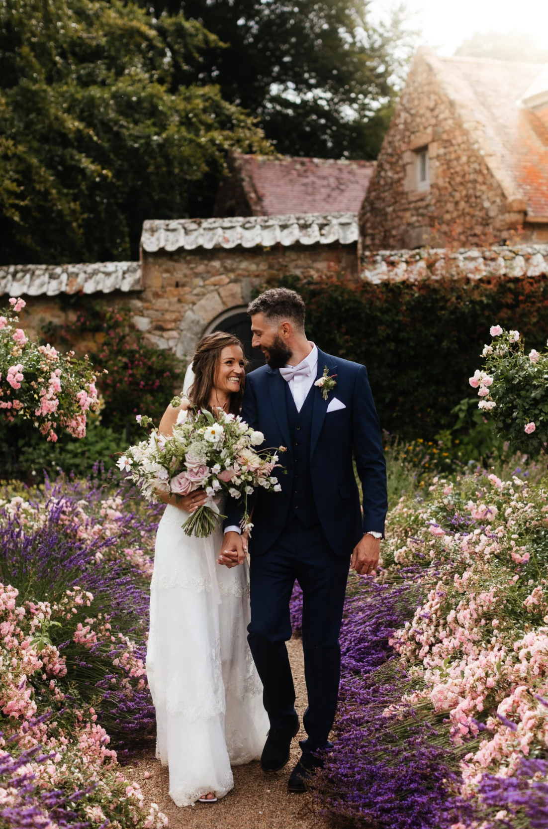 A newlywed couple holding hands and smiling at each other in a garden filled with pink and purple flowers, with stone buildings and trees in the background.