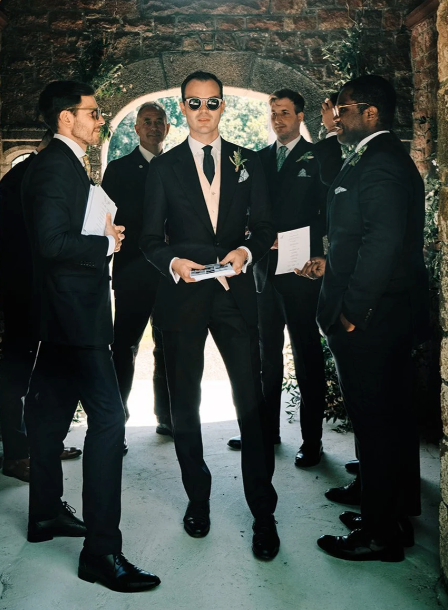 Group of men in suits with boutonnières, some holding papers, standing in a brick archway with sunlight behind them, at a wedding ceremony.