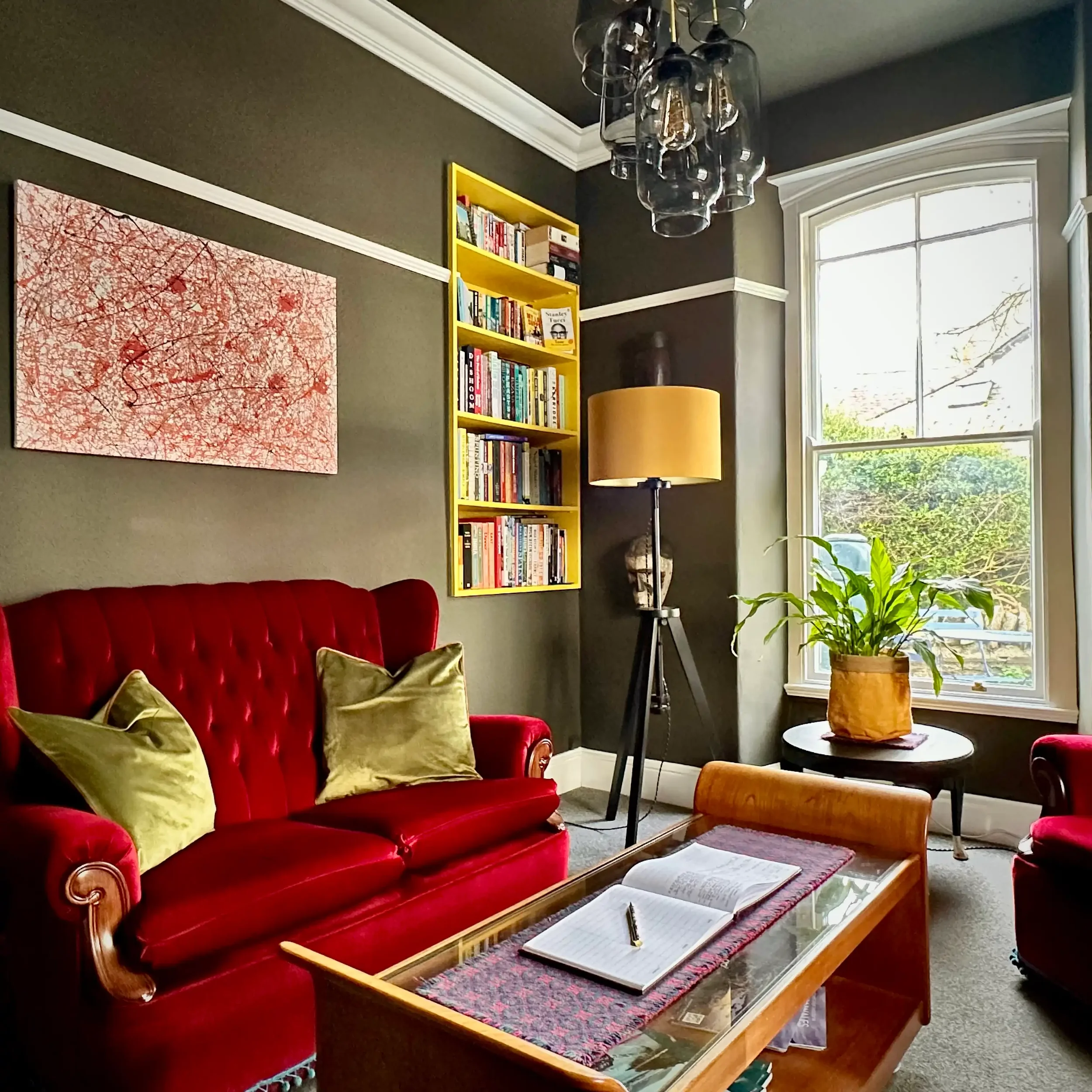 A lounge in a house with dark brown painted walls, a bookshelf and a red sofa with green cushions.