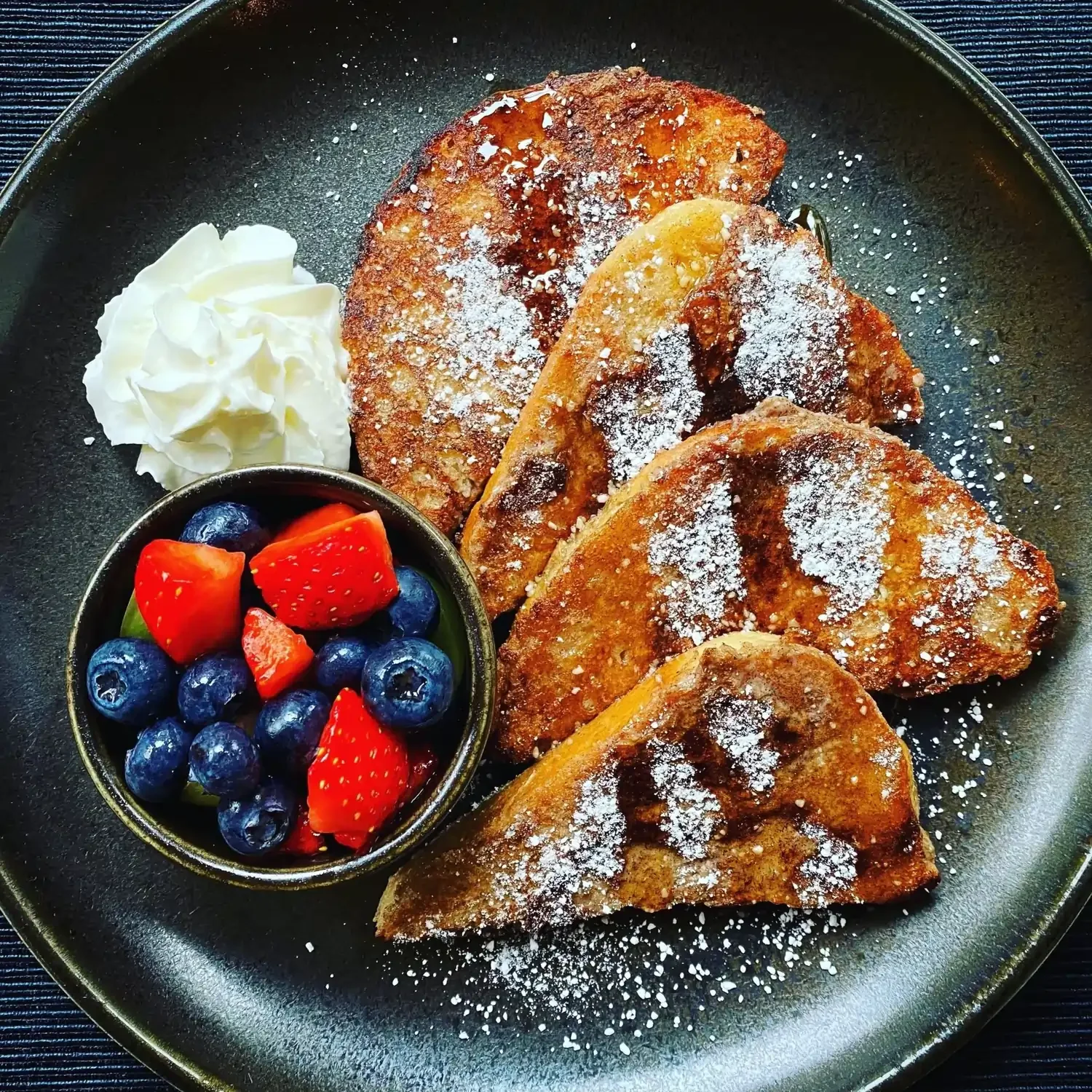 French toast on a black plate with cream and fresh fruit in a small bowl