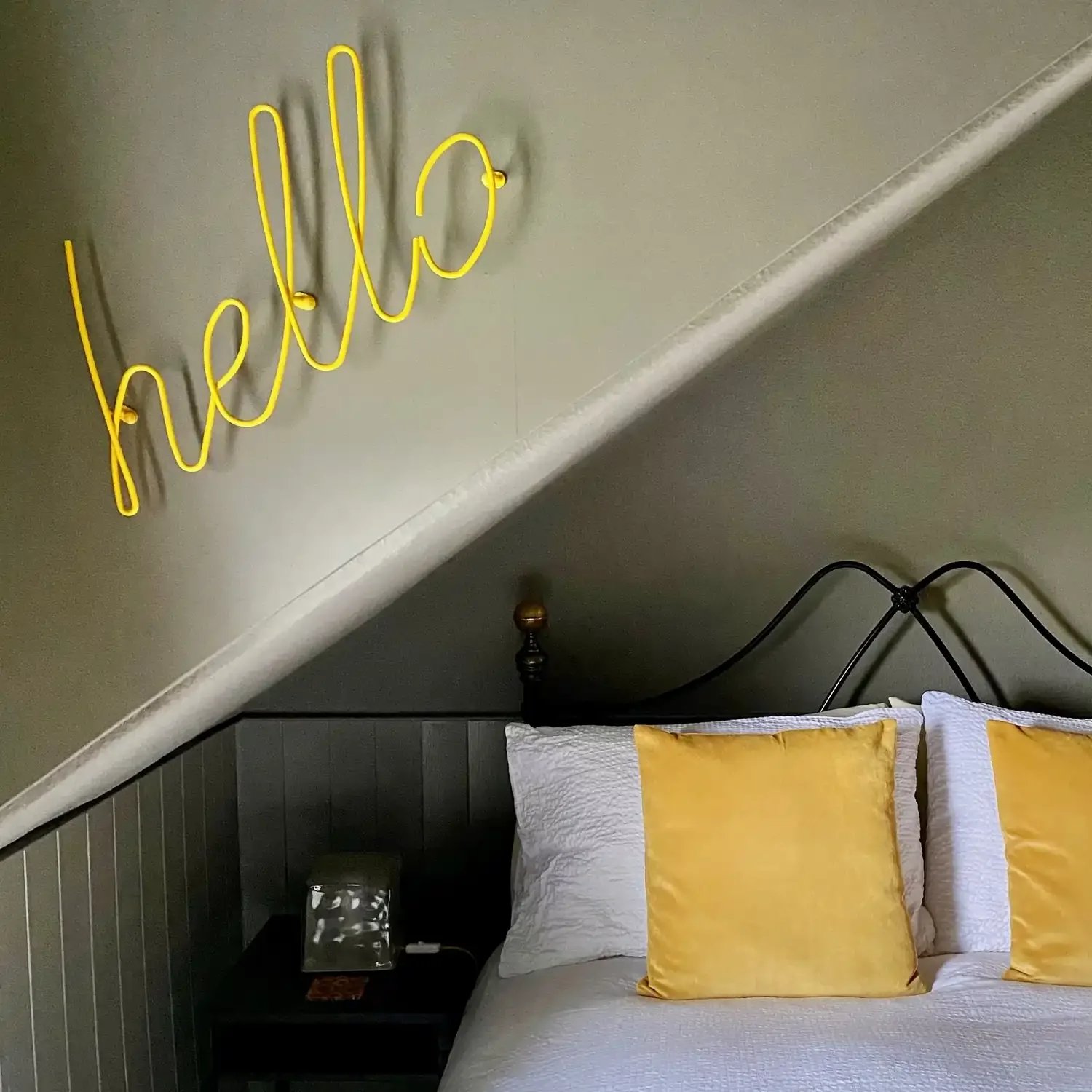 A bedroom in which is a bed with a white duvet cover and yellow cushions, and a sign on the wall saying "hello"