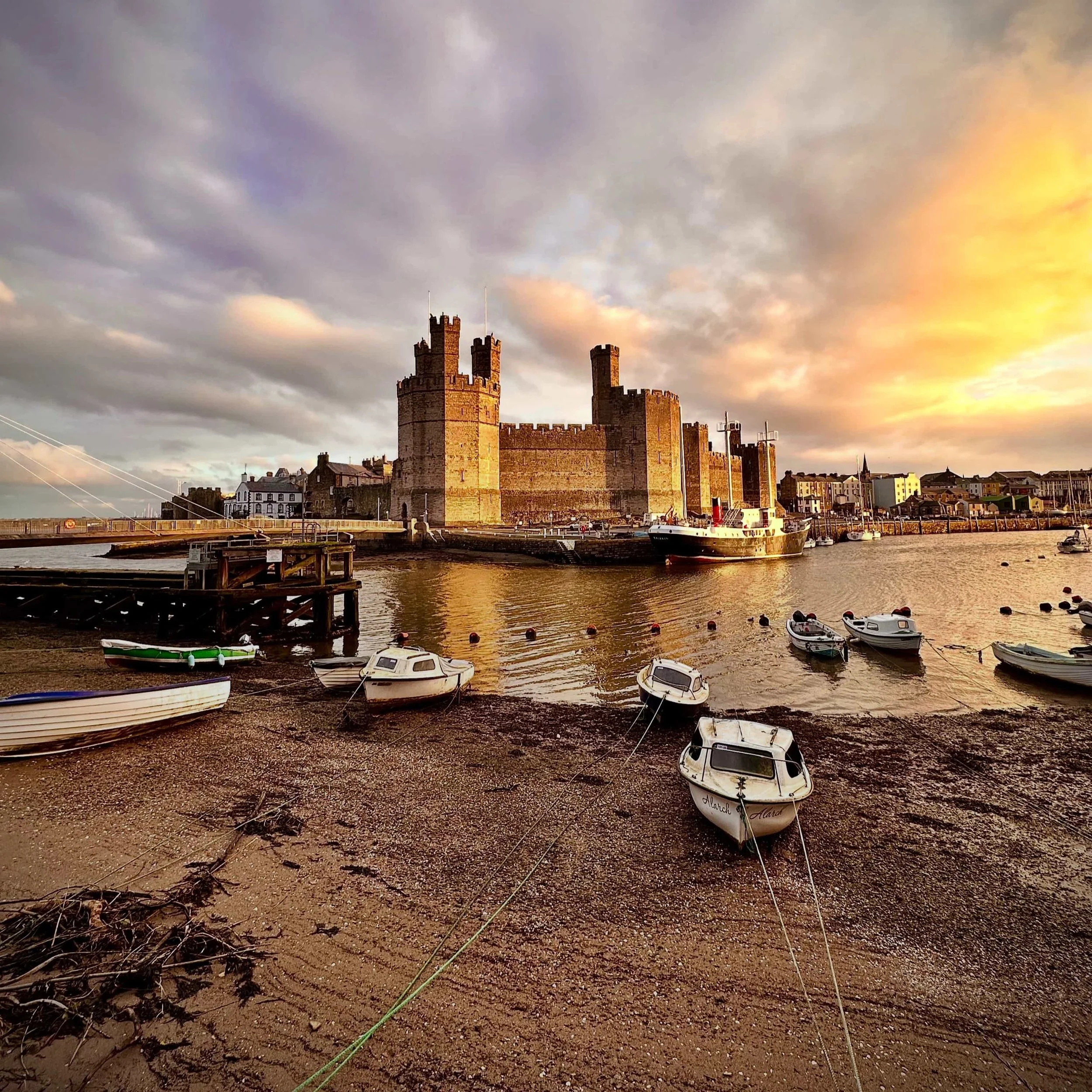 A view of a castle in Wales across a harbour, with boats in the foreground and a sunrise behind it