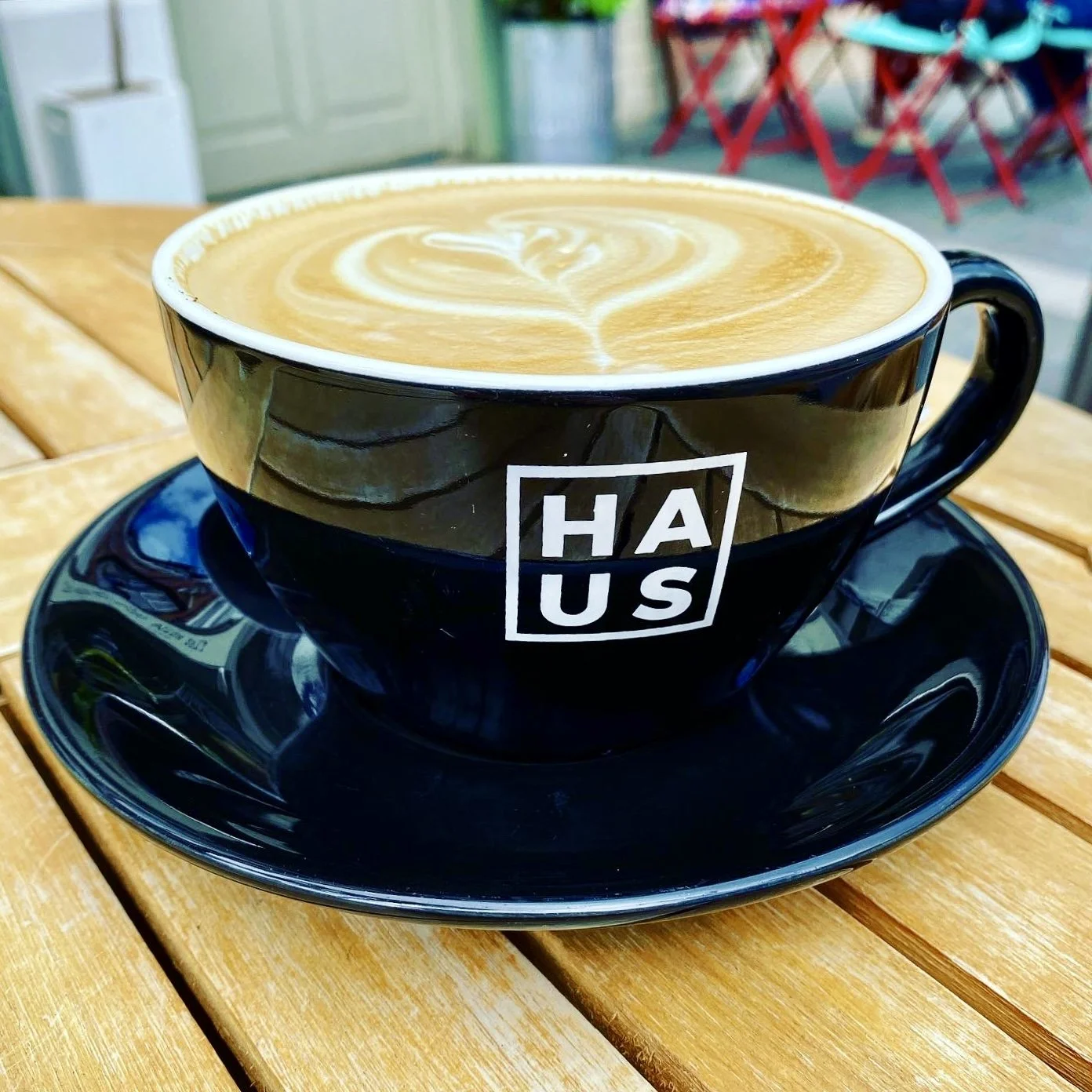 A black coffee cup on a black saucer with the letters HAUS on the front of it
