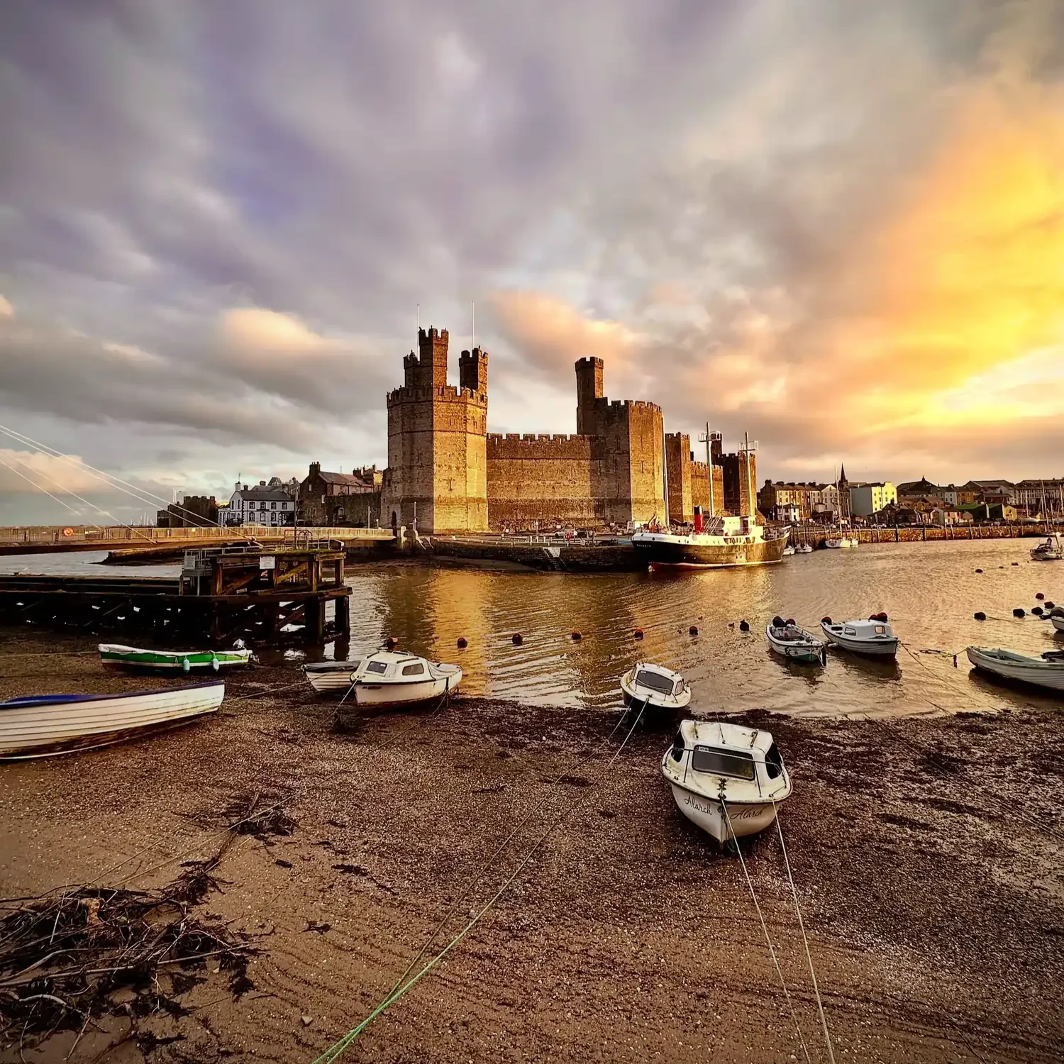 A view of Caernarfon Castle in North Wales at sunrise, with boats in the harbour in the foreground