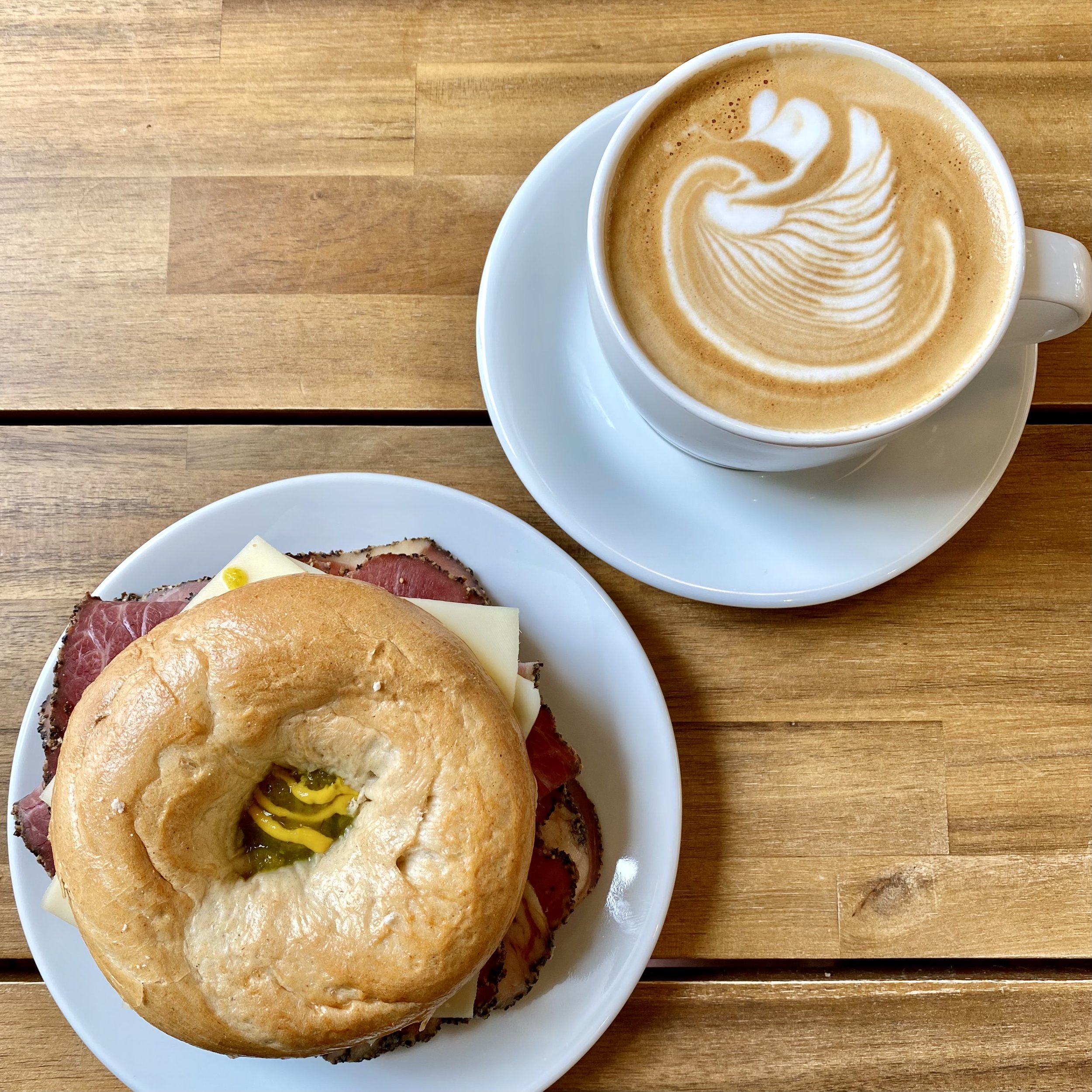 A white cup and saucer containing coffee with latte art on top, next to a white plate on which is a pastrami bagel