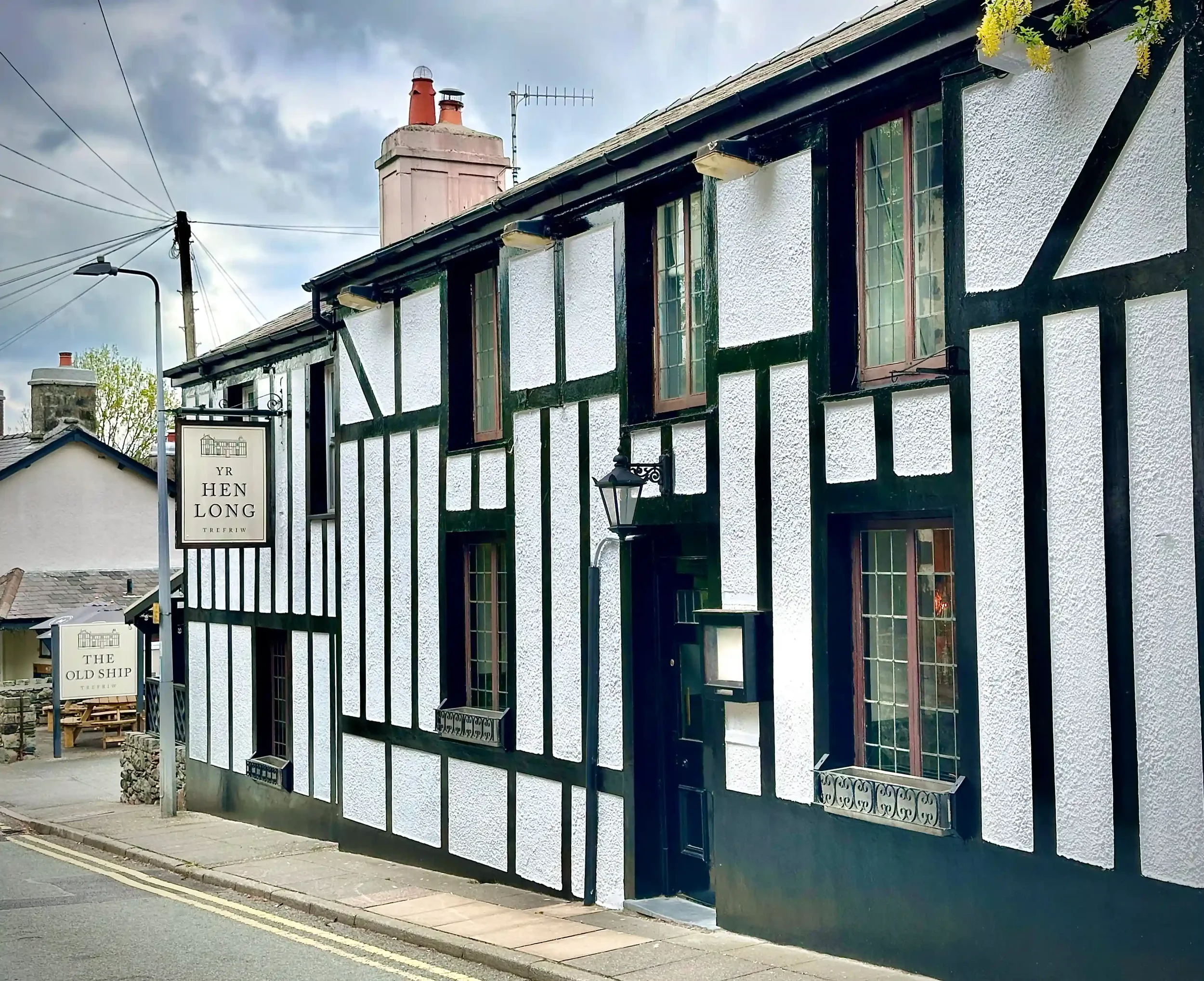 The outside of a village pub in Wales. It is painted black and white and there is a sign saying YR HEN LONG hanging from the wall.