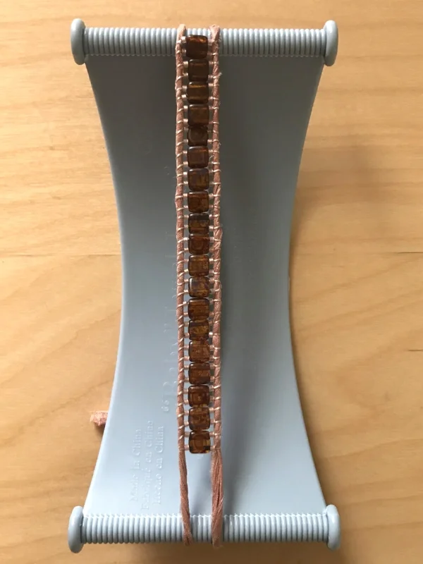 11. Repeat the entire dance by guiding the needle back under the work—left to right—and threading on three new beads. The process will feel not-so-meditative for the first three or four beads while you are still learning, but shortly after, it truly will get easier and you will find yourself getting into a rhythm; completely forgetting about everything else. You will enter a flow of creation, even though you are paying full attention to what you're doing. Continue adding beads until your strand is just a bit longer than the entire circumference of your wrist. For me this meant 19 two-hole beads—about 4-1/2" (11cm). If your thread gets too short to work with, tie the thread on one ribbon strand and then thread the remaining thread through one row of beads. Cut the excess. Cut a new length of thread and tie it to the left ribbon strand where your work currently ends. Proceed as normal. After you've added a few beads, thread the tail through a row or two of beads and cut off the excess. Do the same for the tail at the beginning of the bracelet.