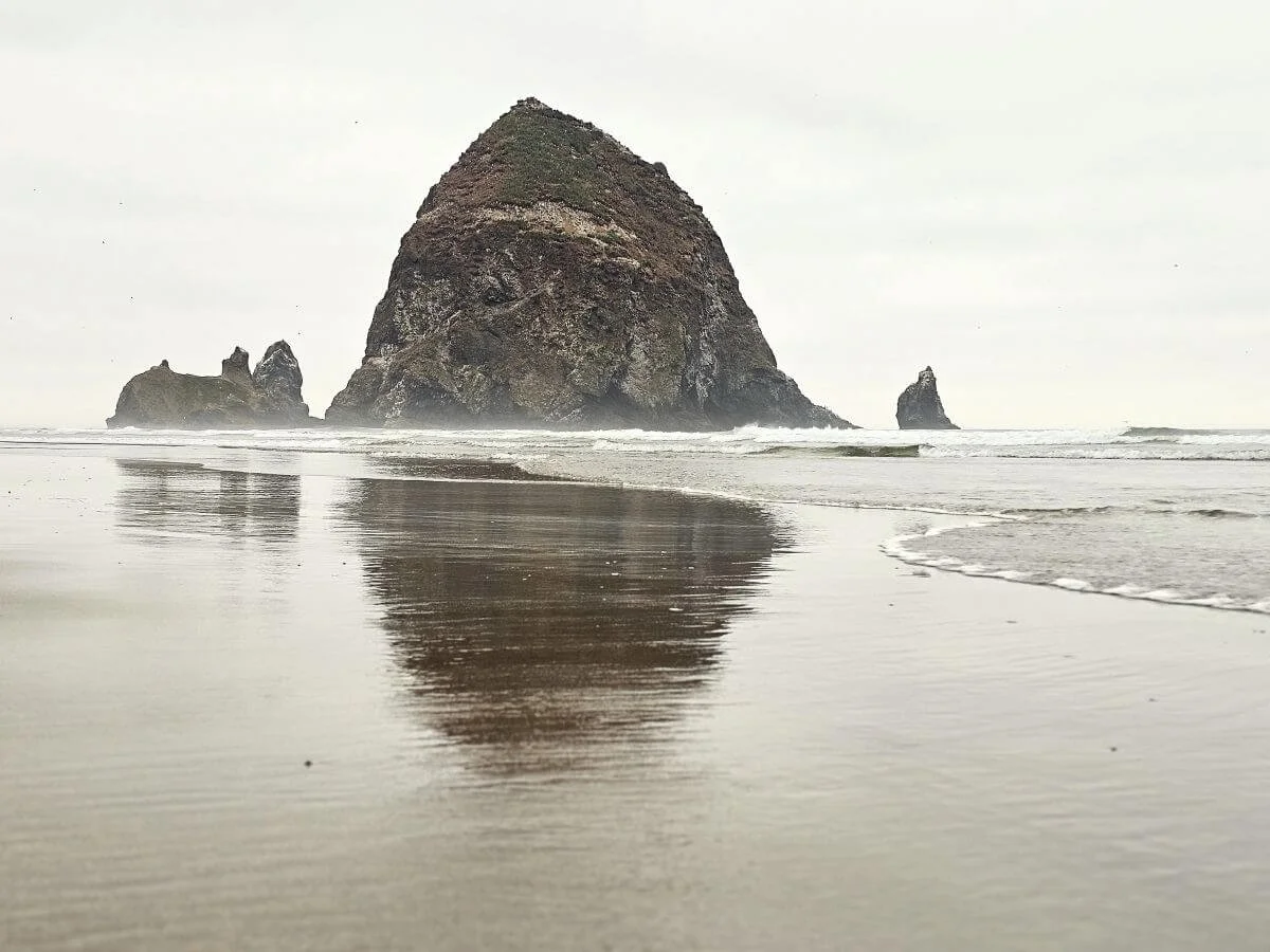 A huge rock in the ocean reflects on the wet sand at Cannon Beach Oregon