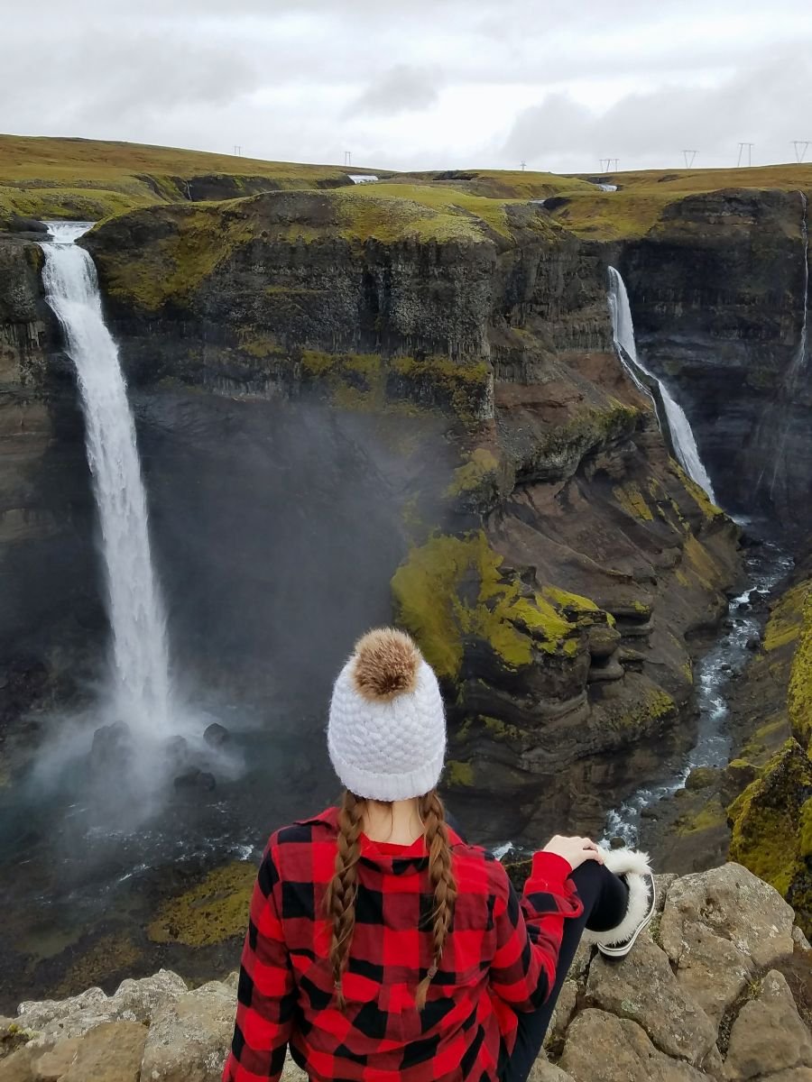 A woman in red plaid sits on the edge of a cliff overlooking Haifoss in Iceland