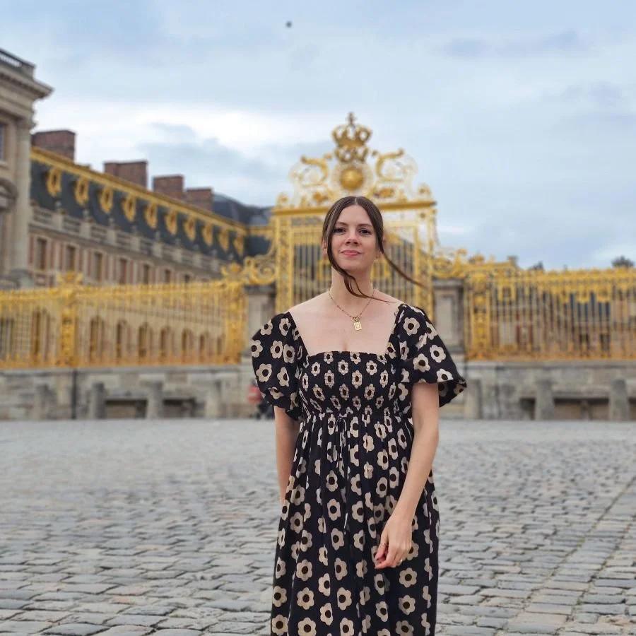 A woman in a floral dress stands in front of a gold gate at a palace