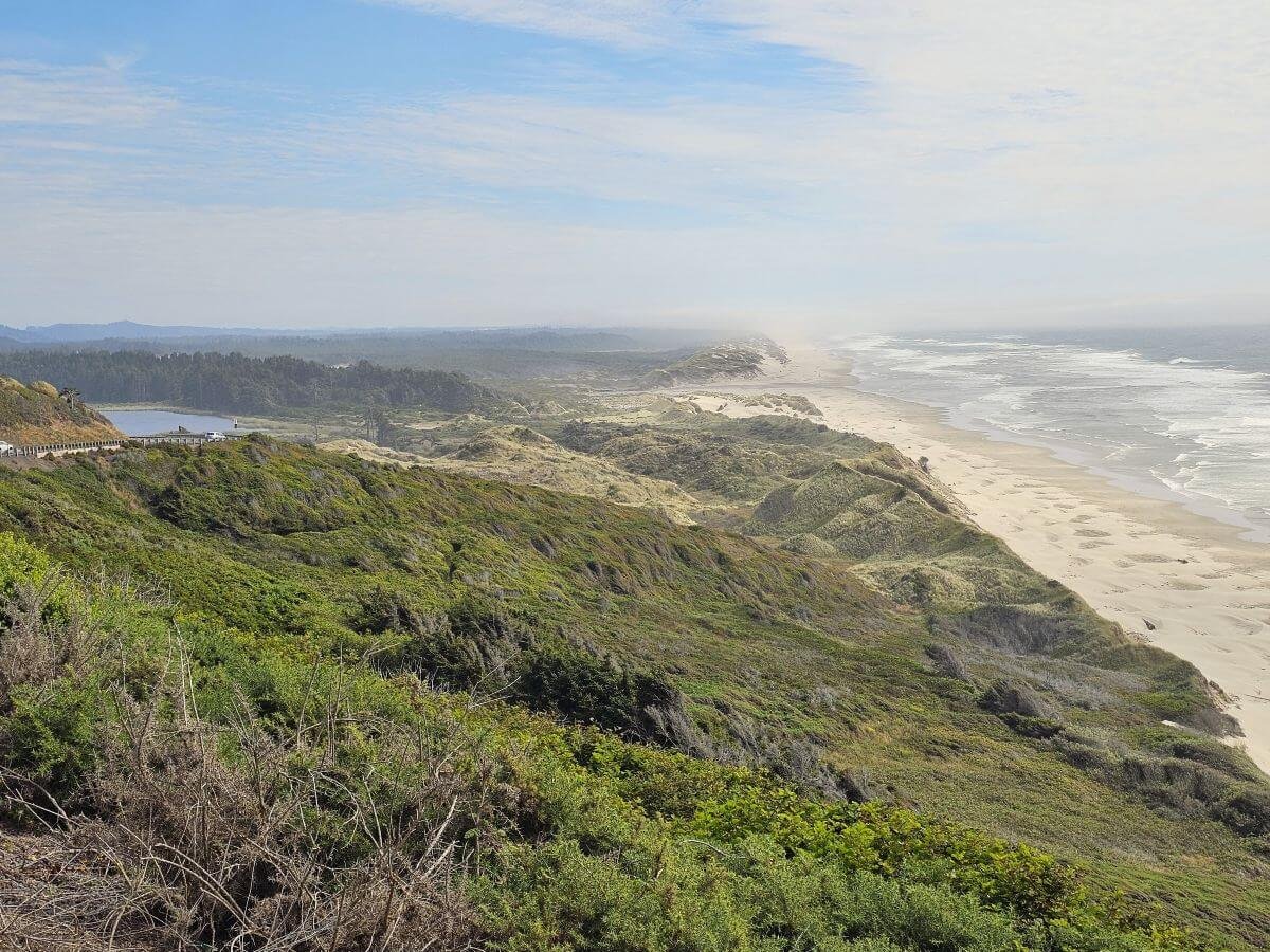 An elevated view of the Oregon coast, with a sandy beach below rolling hills to the right and a road to the left.
