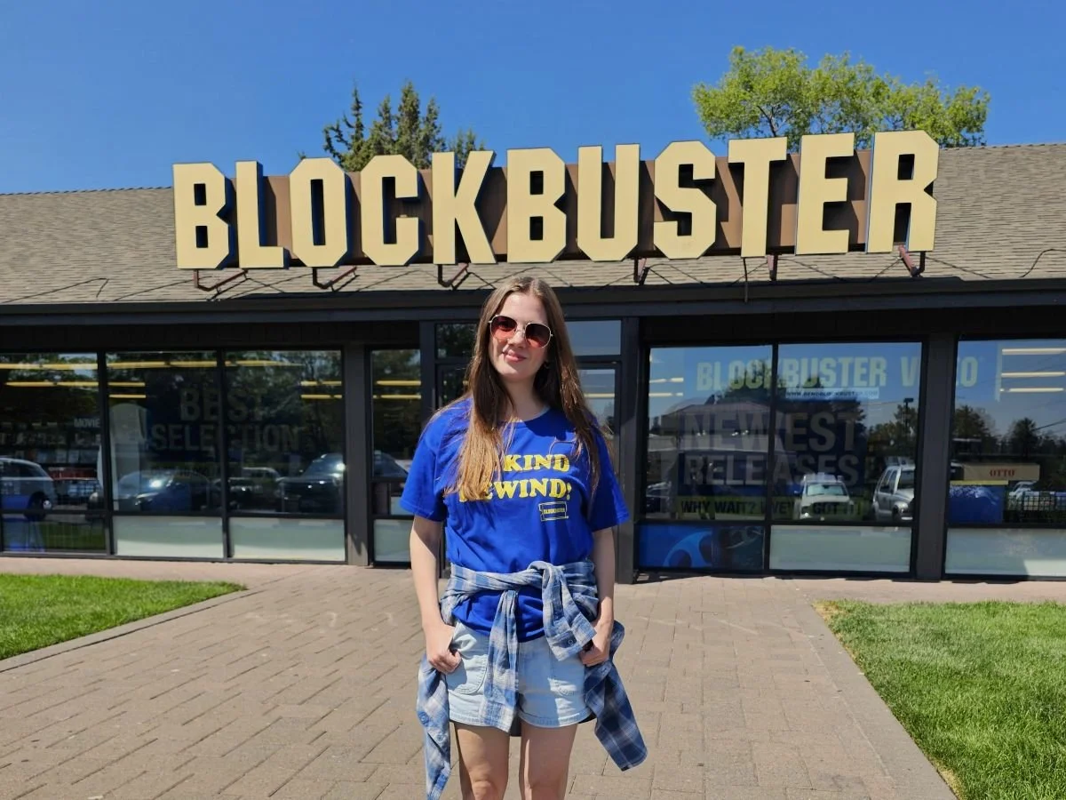 A woman stands in front of a store with giant Blockbuster letters wearing a blue and yellow shirt that says "be kind, rewind."