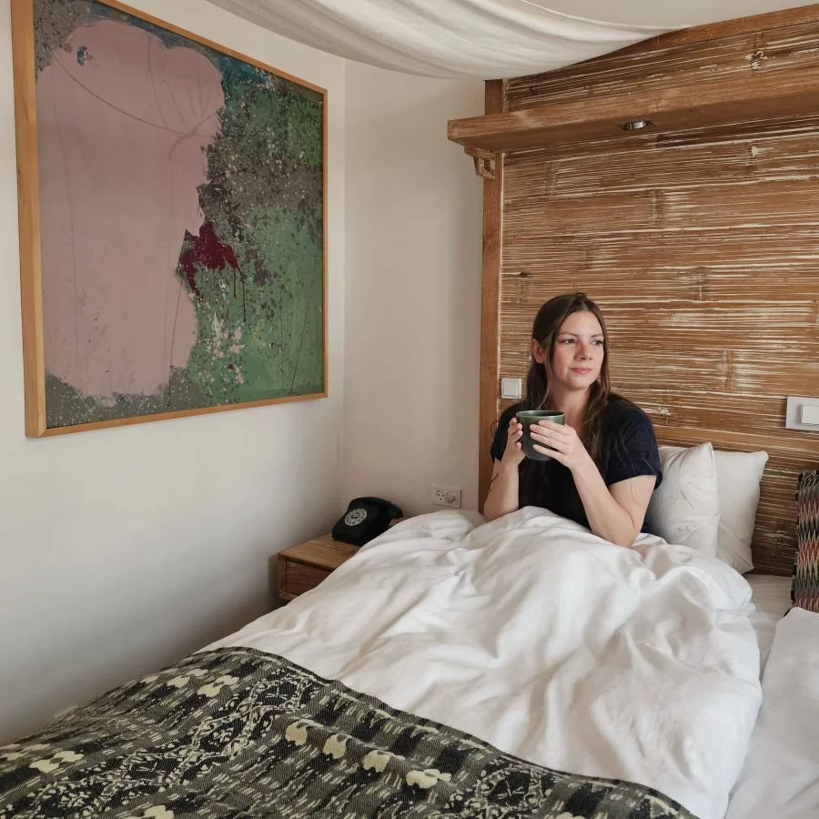 A woman in a dark blue shirt sits in a bed with the white covers pulled up over her lap holding a green mug at a hotel in Copenhagen.