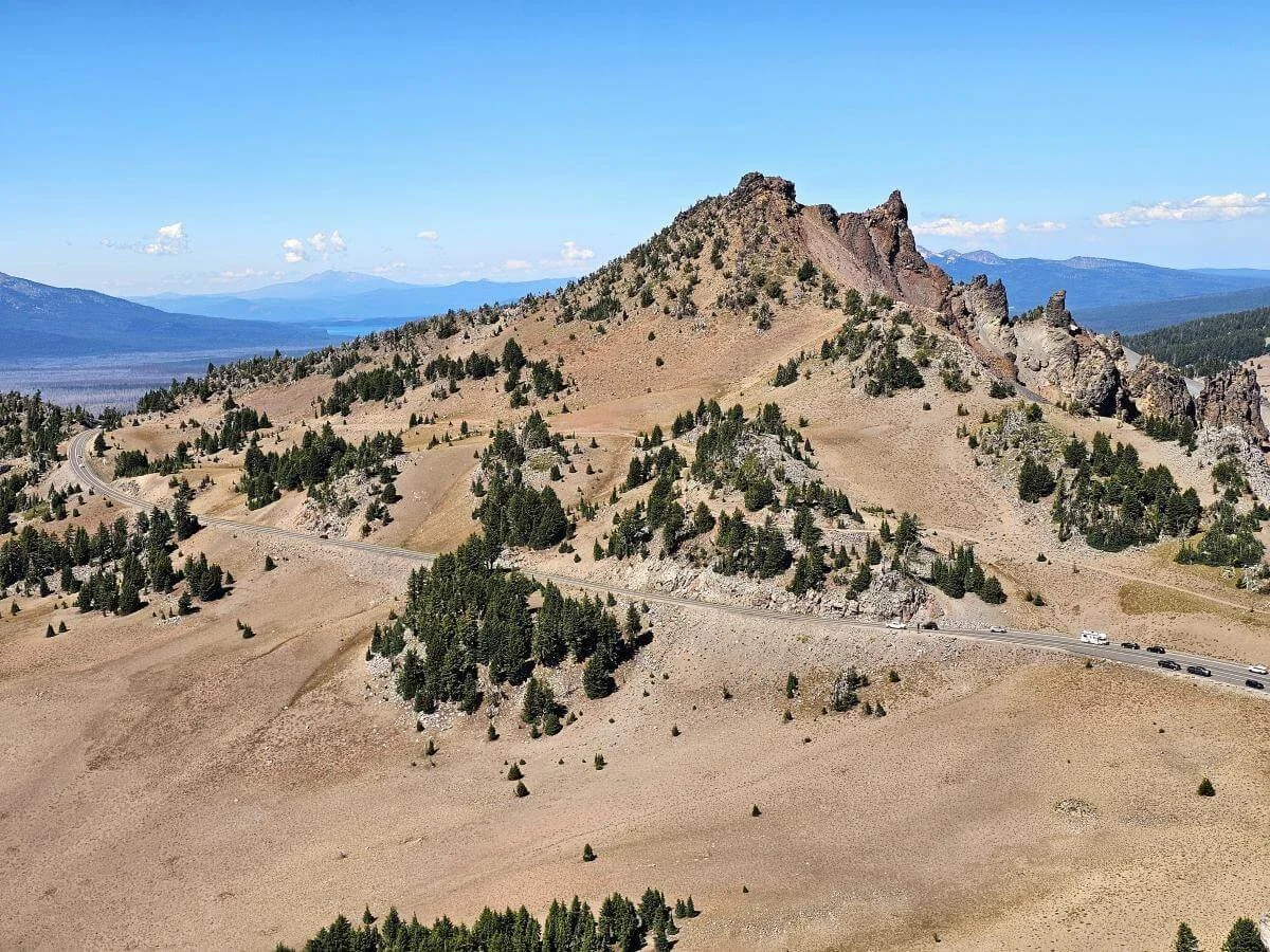 A road runs through a sandy brown volcanic landscape dotted with trees at Crater Lake National Park