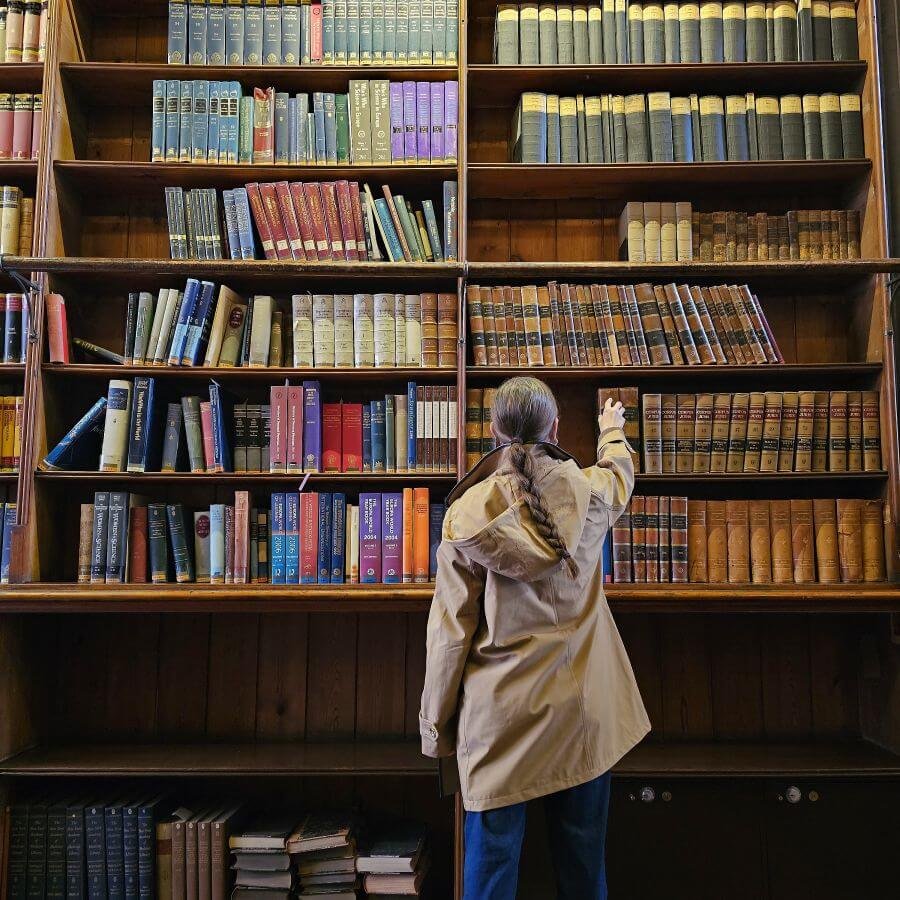 A woman in a trench coat stands with her back to the camera reaching for a book on the tall wall of bookshelves in front of her.
