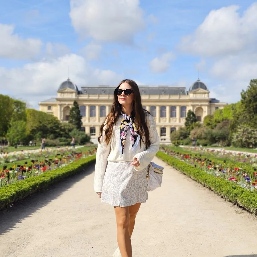A woman in a white skirt and sweater with a scarf around her neck walks toward the camera in a garden with rows of flowers on either side and a building behind her.