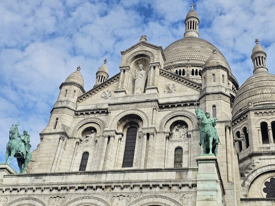 The top half of the white Sacre-Coeur church with green statues of men on horses and a dome at the top under a blue sky with clouds
