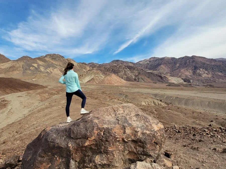 A woman in blue hiking clothes stands on a rock looking out at a landscape of colorful rocky hills.