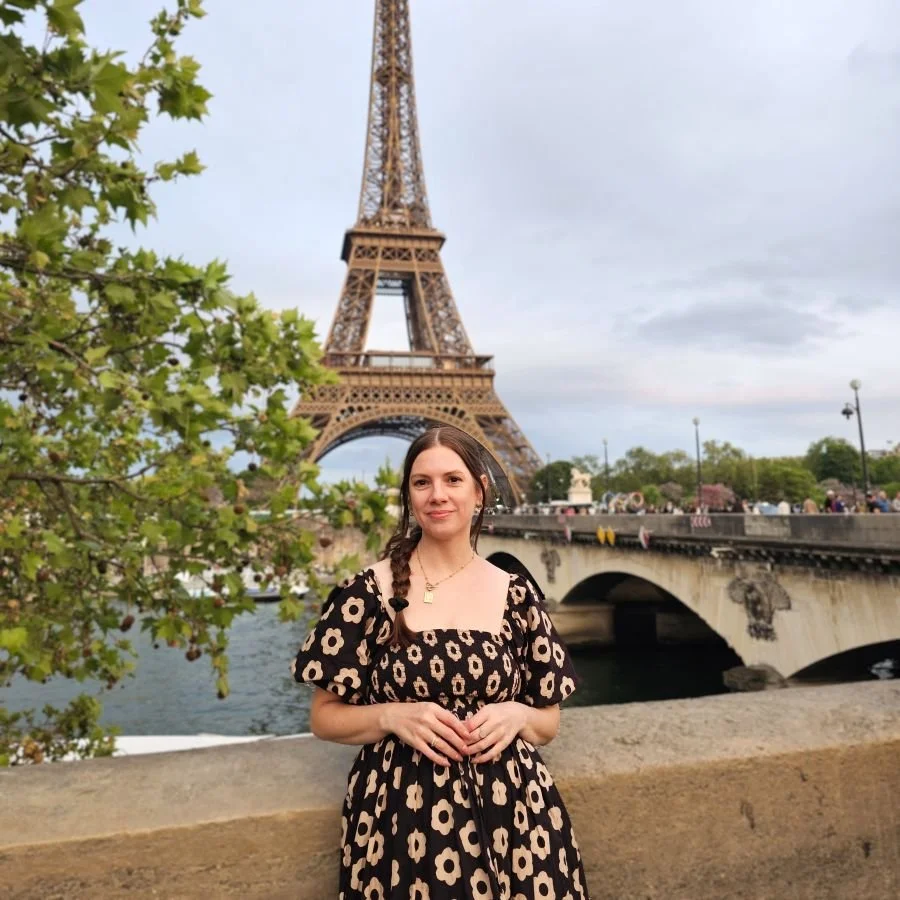 A woman in a floral dress stands with her back to the water and the Eiffel Tower
