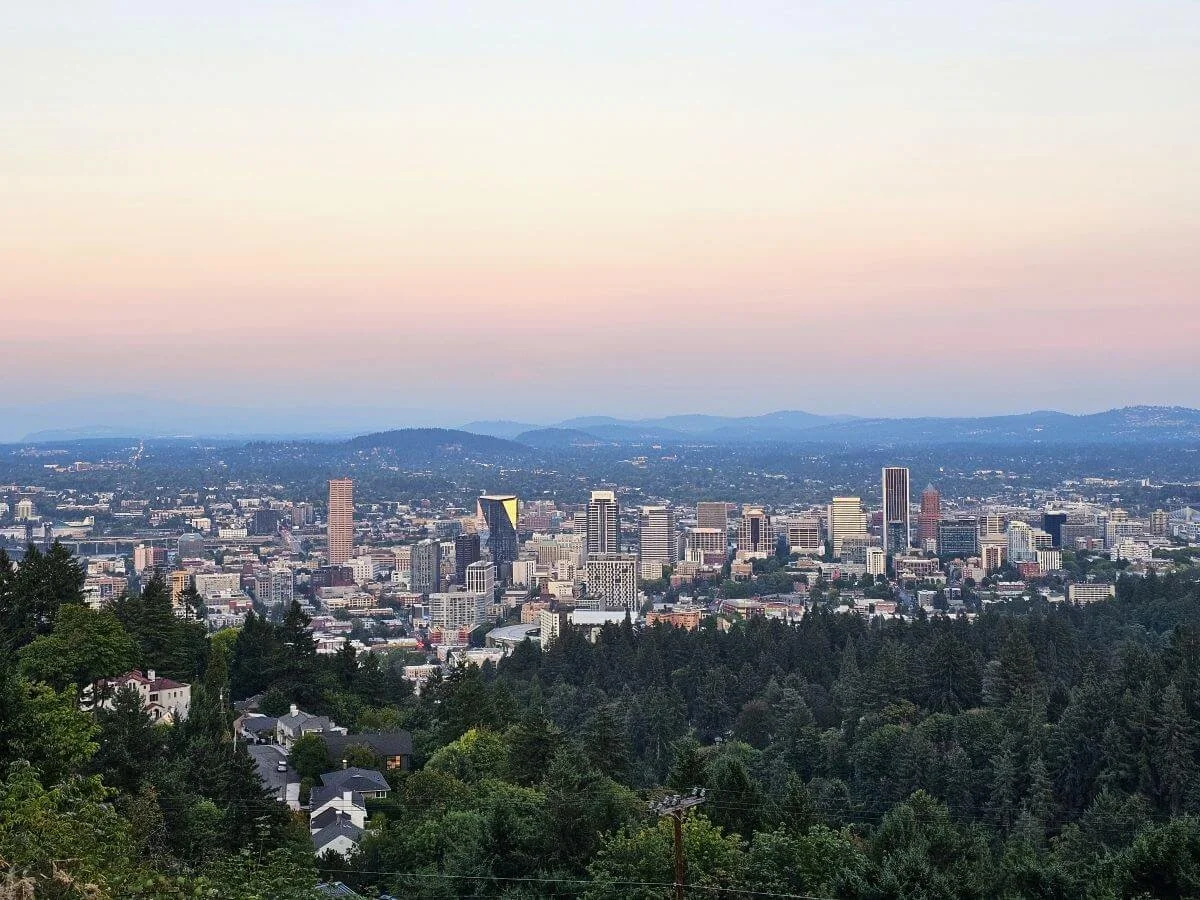 Sweeping overlook of the Portland downtown skyline with mountains behind it and a sunset sky in shades of muted pink and orange