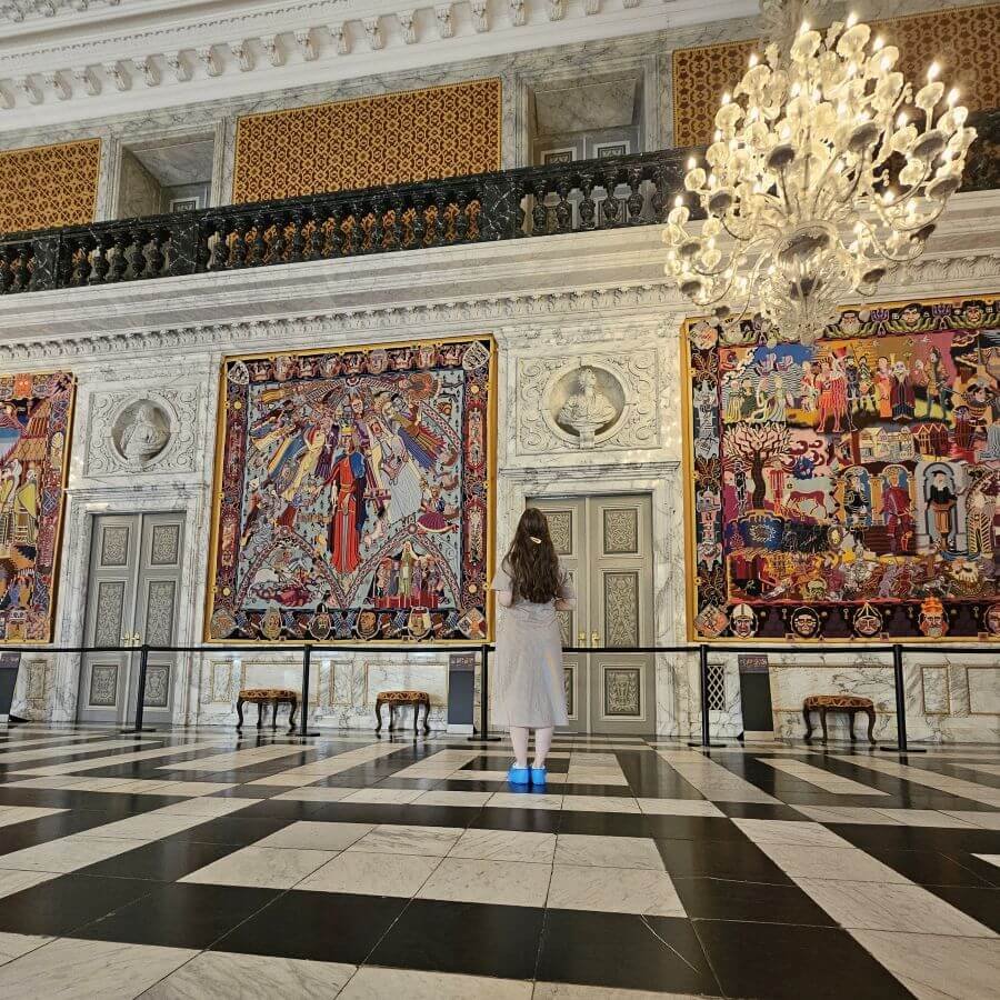 A woman in a dress and blue shoe covers stands on a black and white tile floor looking at colorful tapestries hanging on the wall in Christiansborg Palace