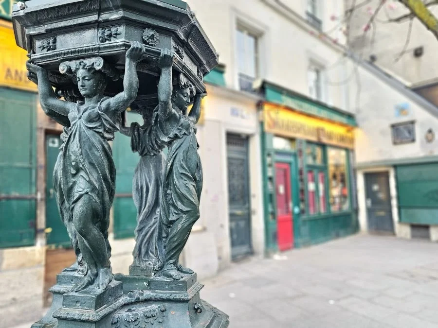 A statue of women lifting an object over their heads with the green storefront of Shakespeare and Company behind it.
