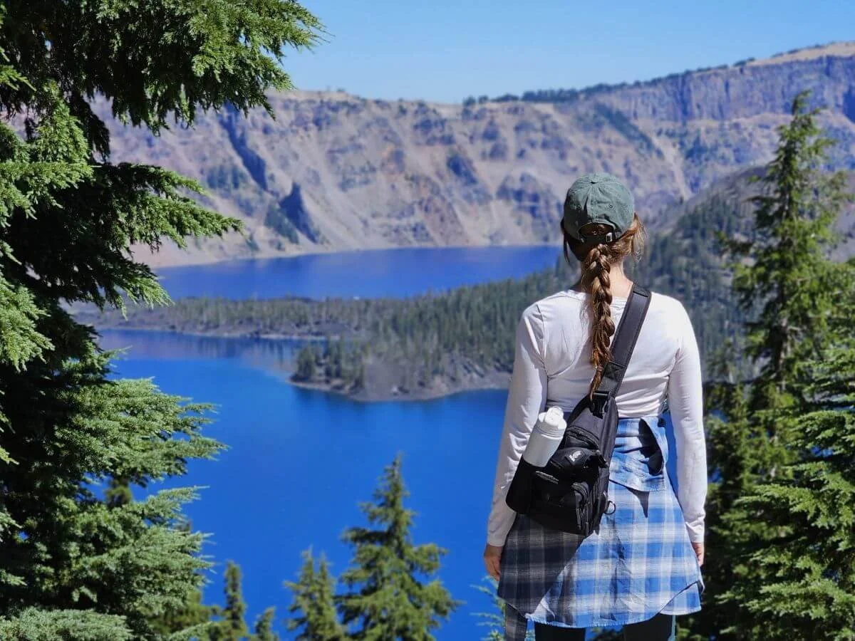 A woman with a blue plaid shirt around her waist stands overlooking a massive blue lake far below