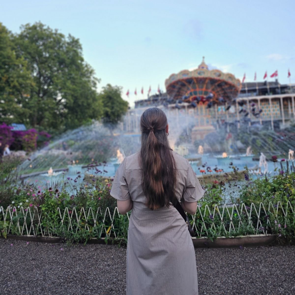 A solo woman traveler with brown hair stands with her back to the camera facing a water fountain at Tivoli Gardens in Copenhagen
