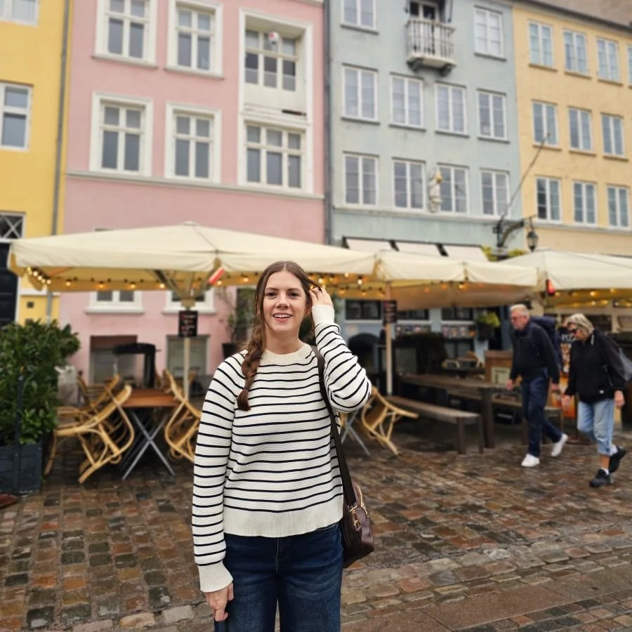 A woman in a striped sweater stands in front of a pink building with tables under an umbrella in front of it in the background.
