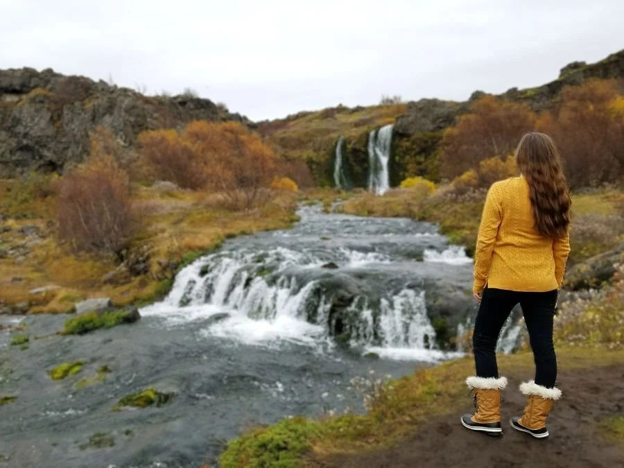 A woman in a yellow sweater stands next to a stream and waterfall