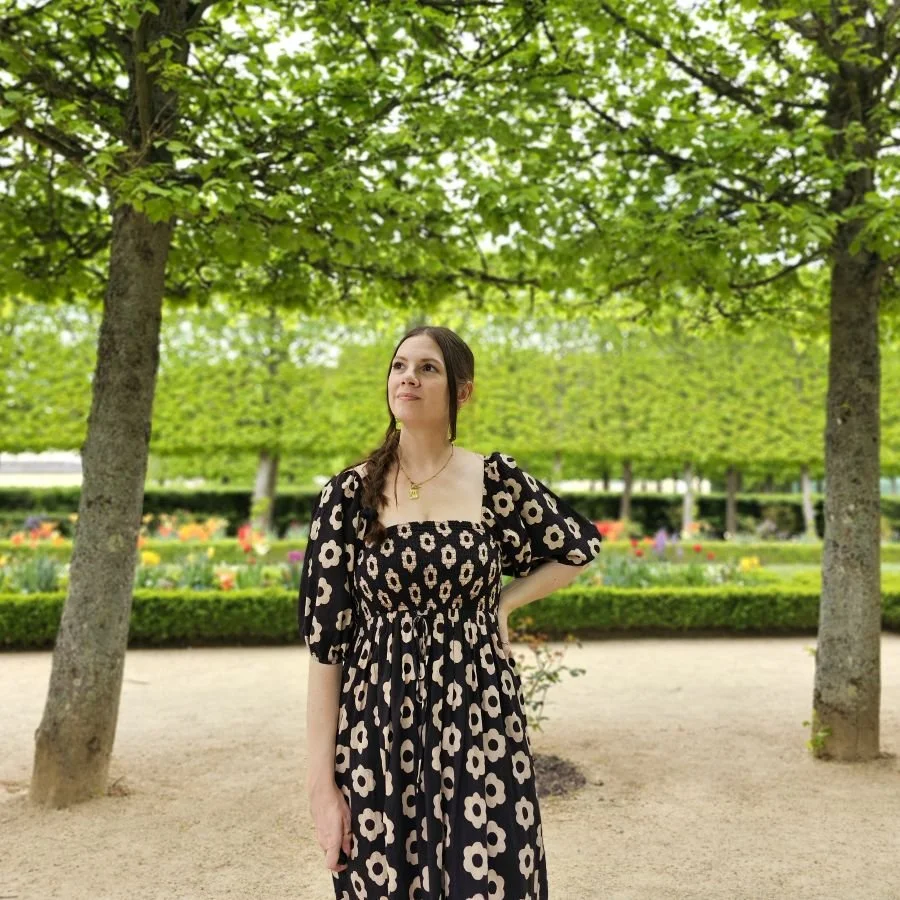 A woman in a floral black dress stands under green trees with a garden behind her in Paris