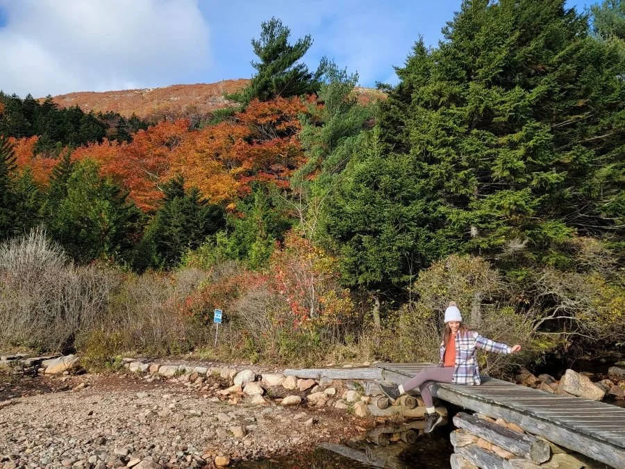 A woman in hiking clothes and a jacket sits on a small wooden bridge with fall colored trees behind her on a solo trip to Acadia National Park.