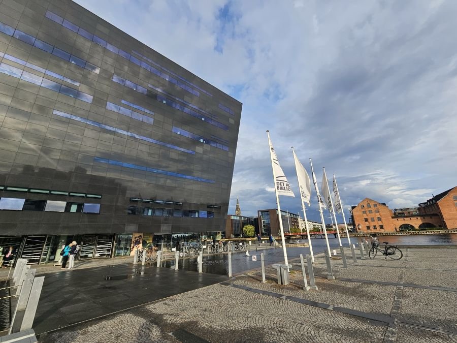 A black angular building on the waterfront with flags flying in front under a cloudy blue sky.