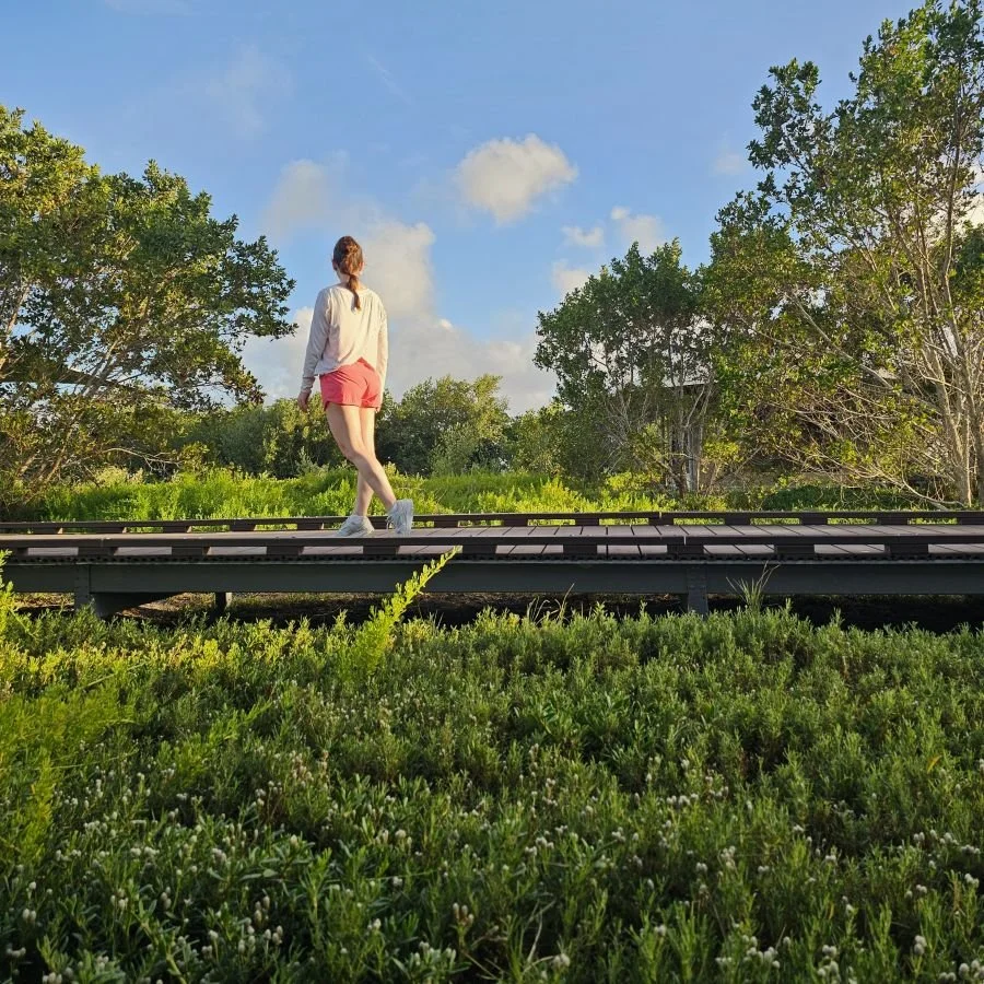 A woman walks along a boardwalk surrounded by greenery