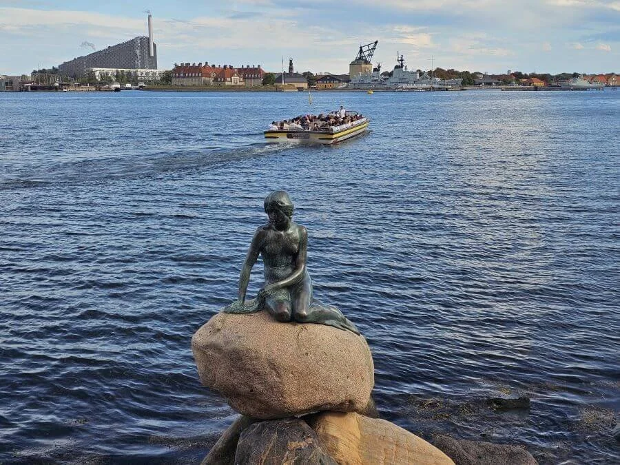 A greening bronze statue of the little mermaid perched atop a rock in the water with a flat boat full of people floating behind it.