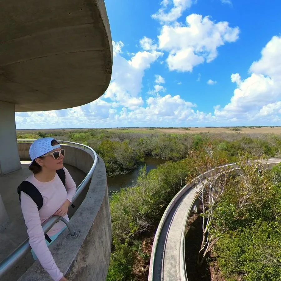 A woman in a white hat and pink shirt stands at the railing of a concrete lookout tower with the swamps of the Everglades below her.