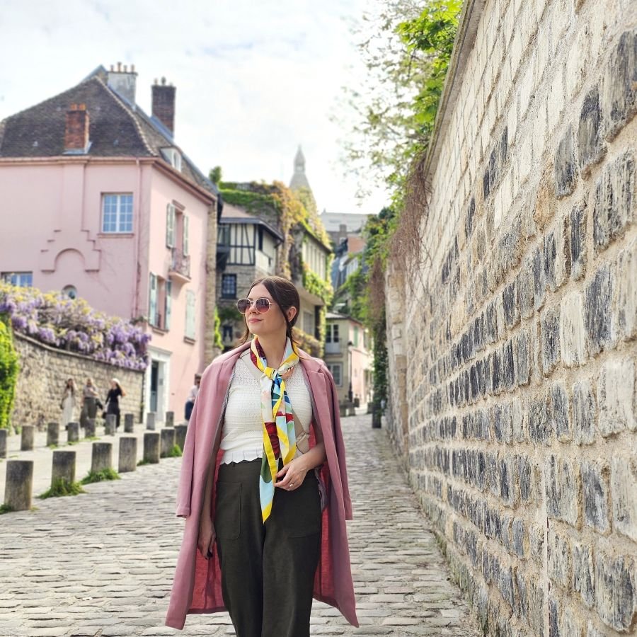 A woman in a pink coat and silk scarf walking down a stone street with a pink building in the background in Paris