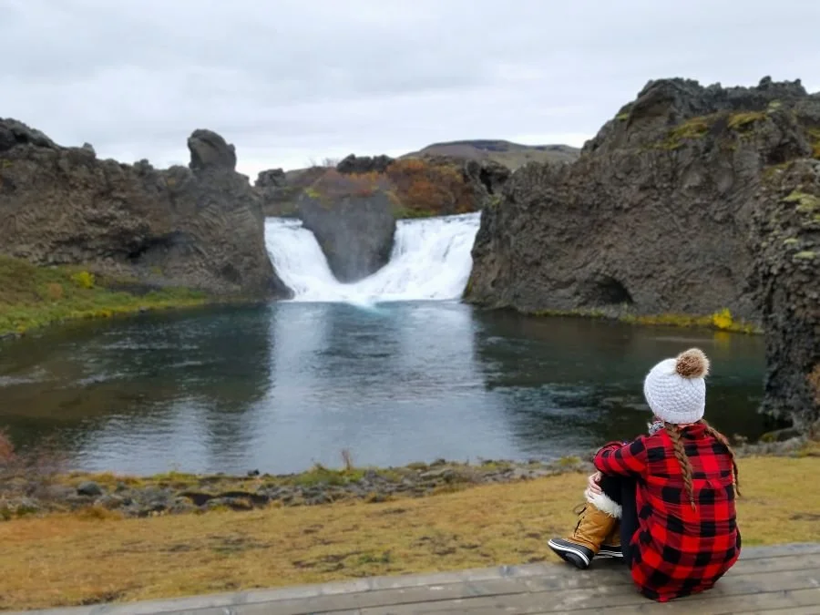 A woman in red sits looking at a waterfall in Iceland
