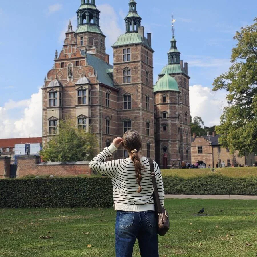 A woman in a striped sweater and braid stands facing away from the camera looking at a small castle under a cloudy blue sky.
