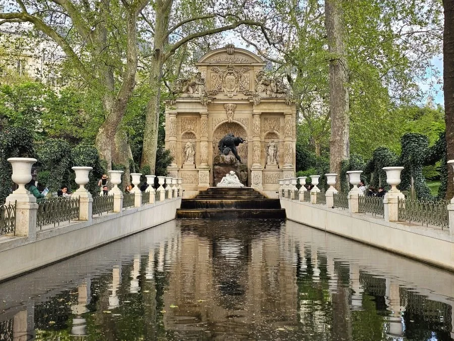 This Paris itinerary 2 days includes the Luxembourg Palace, pictured here reflecting into a pond and fountain in front of it under a blue sky.