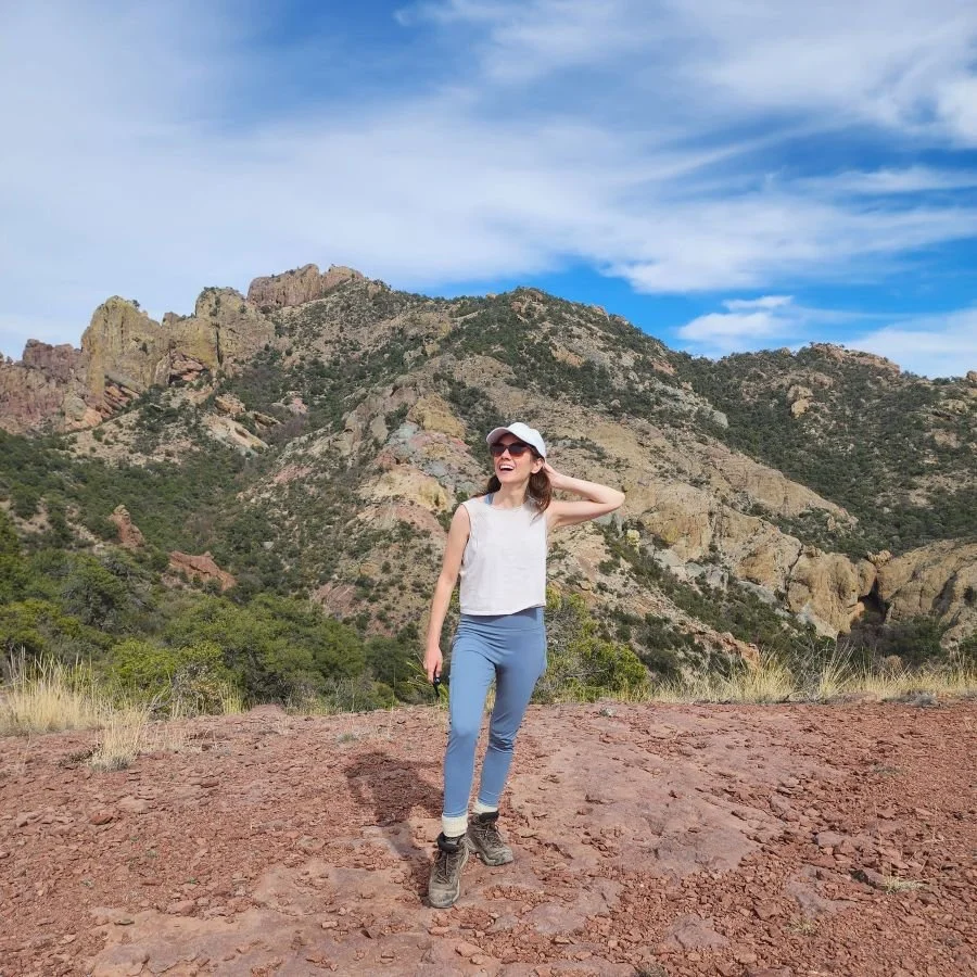 A woman in hiking clothes and a ballcap stands in front of a colorful rock mountain in Big Bend National Park.