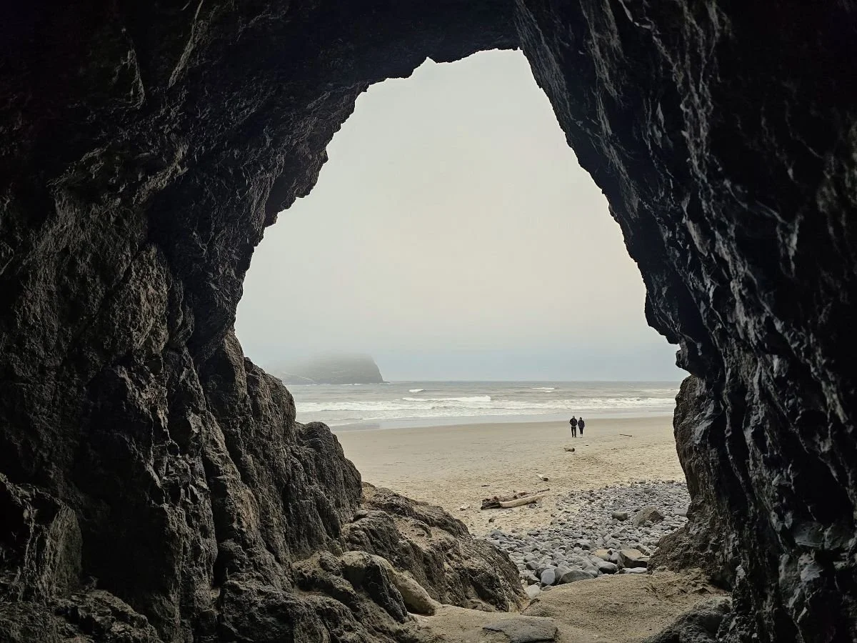A tunnel through a rock formation looks out on a sandy beach in Oregon.