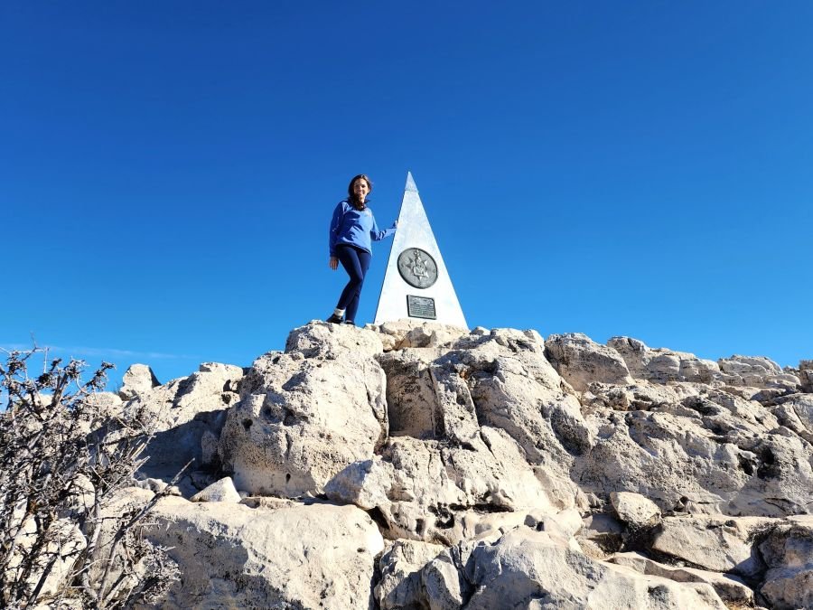 A woman stands on a pile of rocks next to a market signifying a mountain peak at the highest point in Texas on Guadalupe Mountain