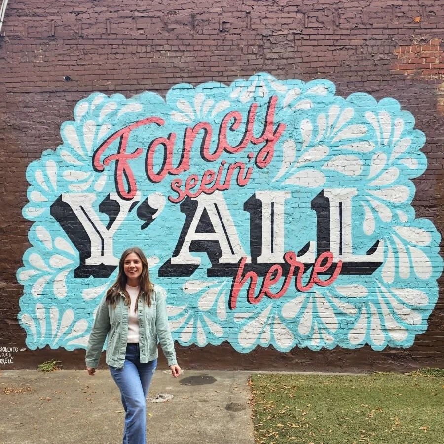 A woman in jeans and a jacket stands in front of a mural in Chattanooga