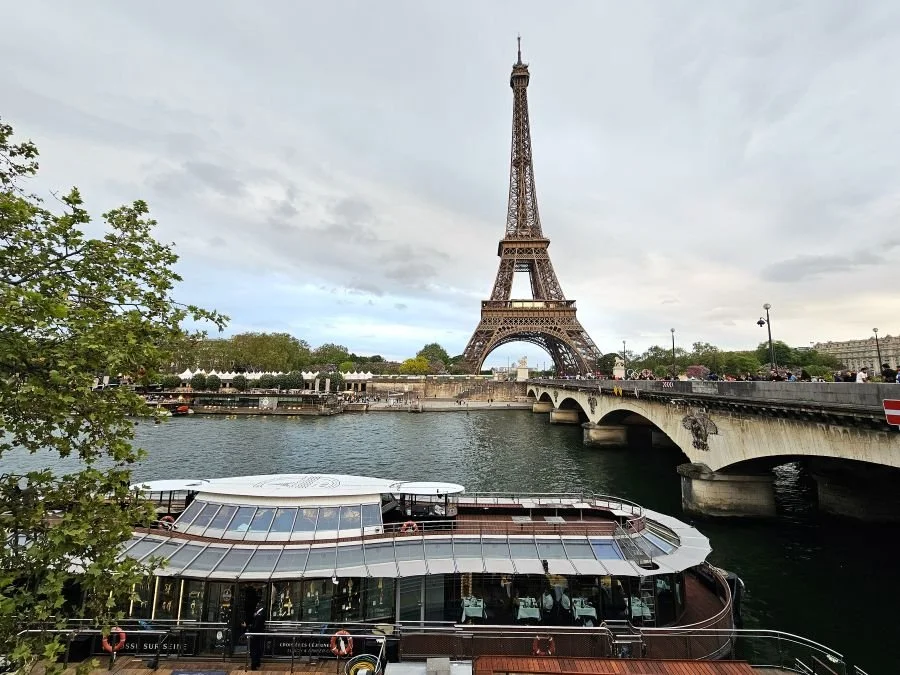 The Eiffel Tower under a cloudy sky with the river full of boats and a bridge below it in Paris