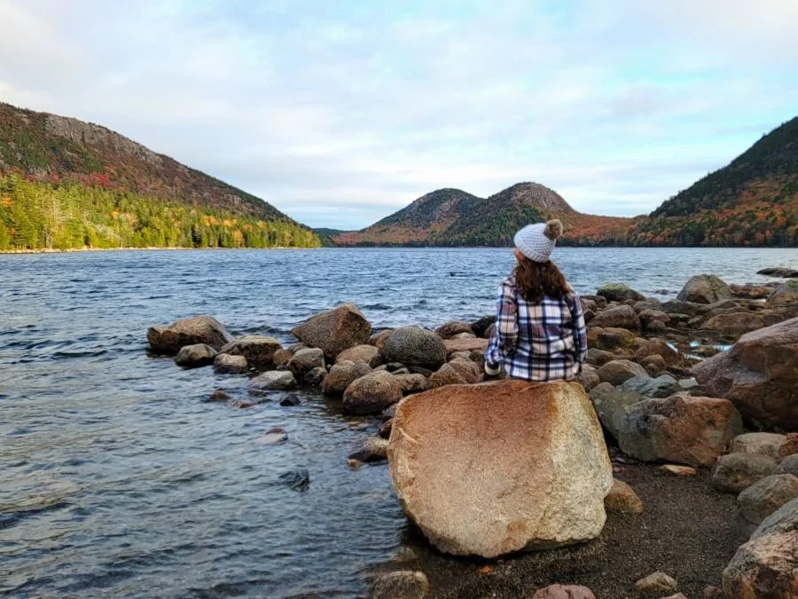 A woman in a plaid jacket and beanie sits on a rock on a lake at Acadia National Park solo.