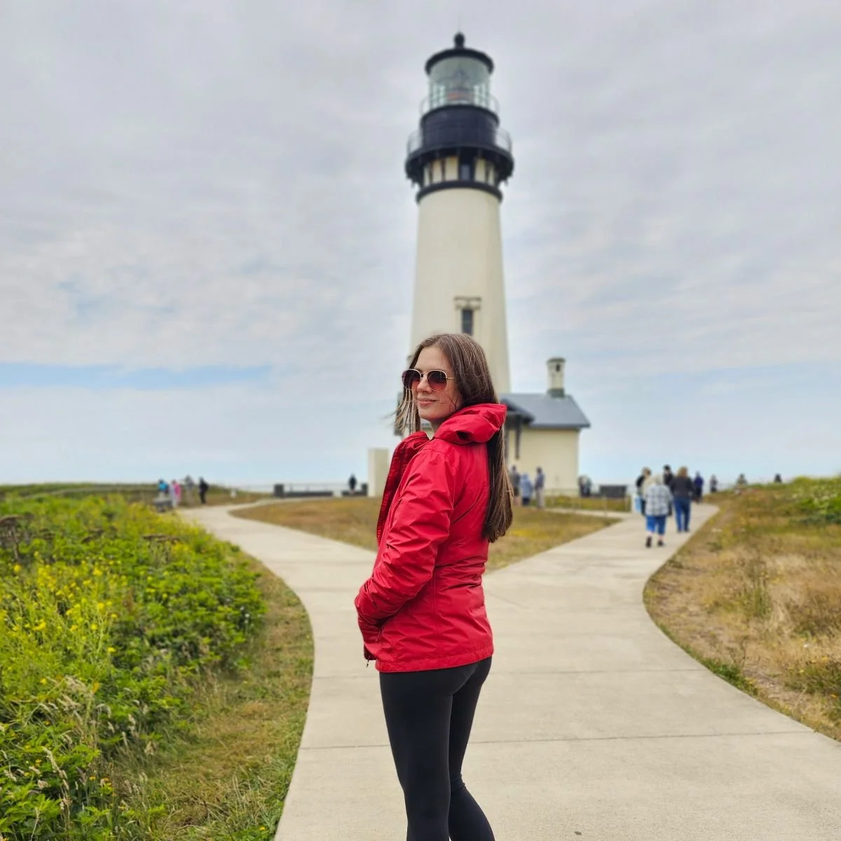 A woman in a red jacket walks toward a lighthouse on the Oregon coast