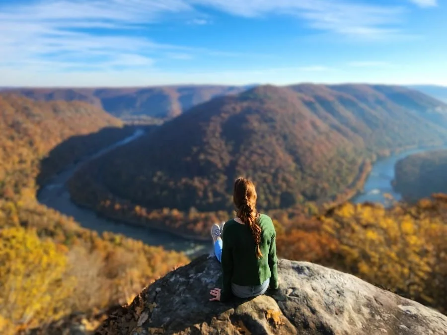 A woman in jeans and a green sweater sits on a rock facing a river bend while solo in New River Gorge National Park